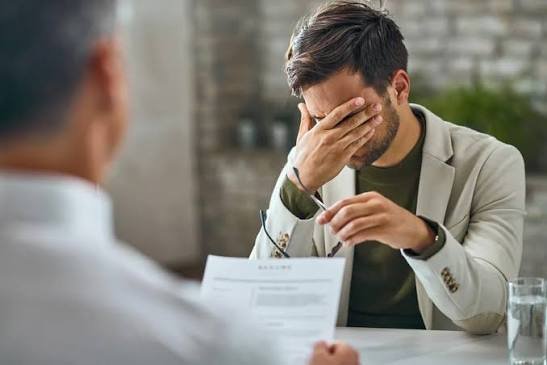 Un homme assis à une table se couvre le visage d’une main, visiblement bouleversé devant un document, tandis qu’une autre personne lui fait face.